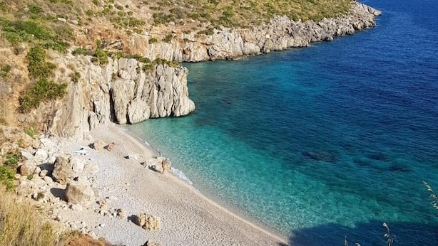 A coastal landscape with a rocky beach, clear blue sea, and a hillside with sparse vegetation. - Cala di Ponente (San Vito Lo Capo)