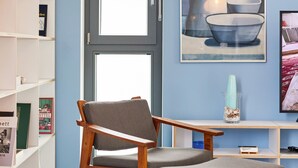Ein Zimmer mit einem Holzstuhl und einem Hocker, einem Bücherregal und einem Bild von Küchenutensilien an der Wand.
