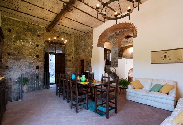 A rustic interior with stone walls, a dining area with wooden chairs and a table, and a white sofa with cushions.