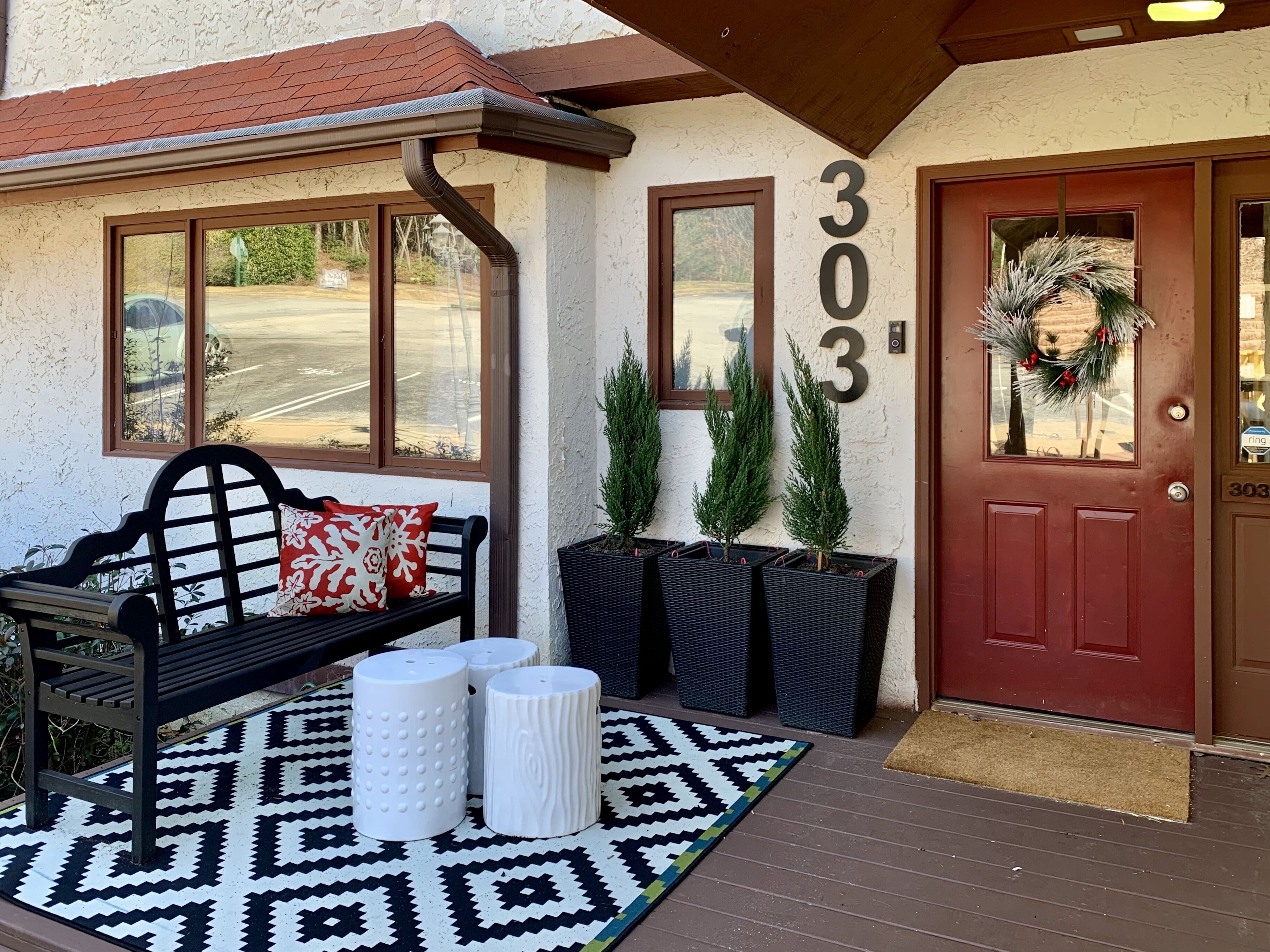 A front porch with a bench, potted plants, a patterned rug, and a red door with the number 303.