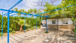 Ein überdachter Außenbereich mit einer Pergola, einer Steinmauer und einem kleinen Gebäude mit vergitterten Fenstern.