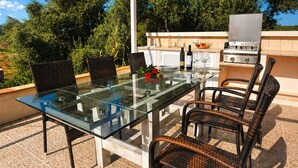 Un área de comedor al aire libre con mesa de vidrio, sillas de mimbre, una parrilla y vista a árboles.