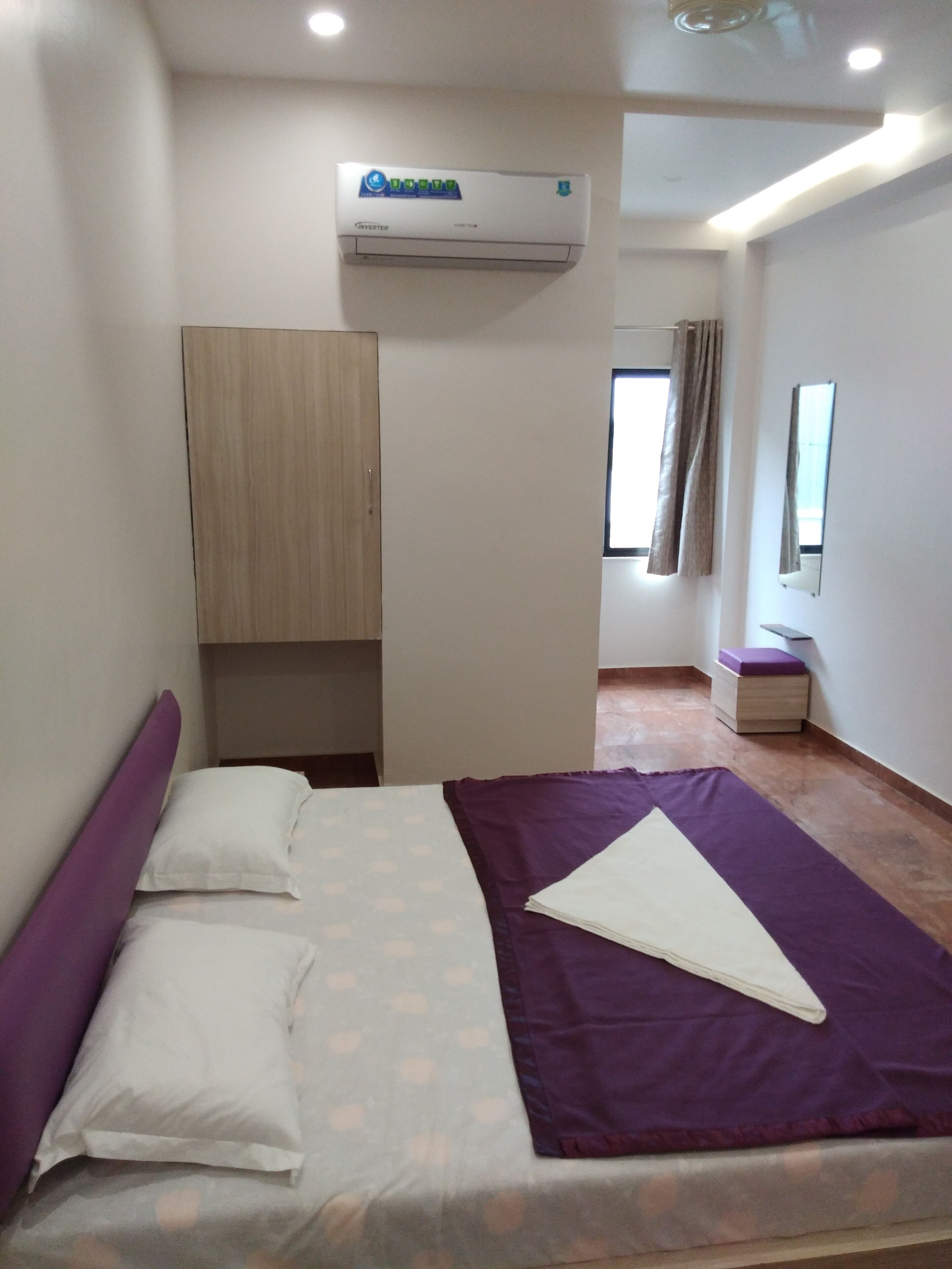 Una habitación pequeña y bien iluminada con cama, armario, aire acondicionado, ventana con cortinas y una mesita.
