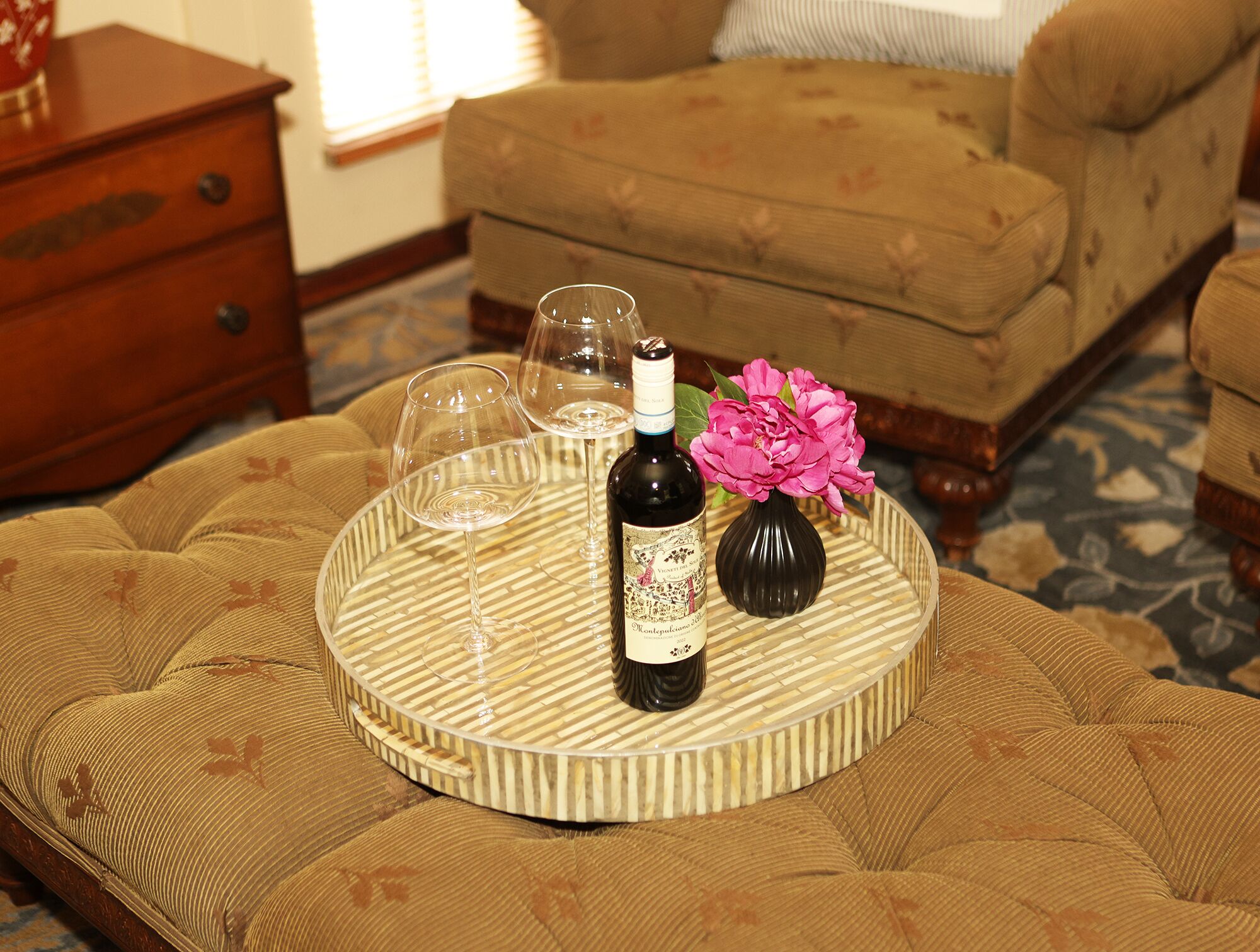 A round tray with a bottle of wine, two glasses, and a vase of flowers on a cushioned ottoman.
