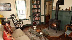 A cozy living room with a sofa, a glass coffee table, a fireplace, a bookshelf, and a window with curtains. - Connecticut Colonial with River and Sunset Views! (East Haddam)