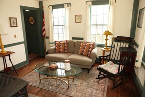 A living room with a beige sofa, patterned cushions, a glass coffee table, a rocking chair, and a patterned rug.