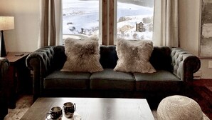 Een gezellige woonkamer met een donkergroene bank, twee zachte kussens, een houten salontafel met twee mokken, en een sneeuwlandschap dat zichtbaar is door grote ramen.