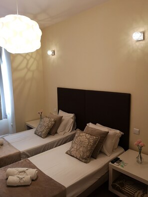 Habitación de hotel con dos camas individuales, un ventanal grande, una lámpara colgante y una mesita con un jarrón de flores.