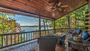 A covered deck with wicker furniture, a table, and a view of a lake surrounded by trees. - Lodge On The Lake- Pet Friendly | Waterfront w/ Dock | Beautiful Views (Blue Ridge)