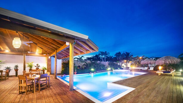 Una terraza de madera con piscina, mobiliario exterior y un espacio cubierto con ventilador de techo.