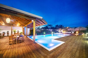 Una terraza de madera con piscina, mobiliario exterior y un espacio cubierto con ventilador de techo.