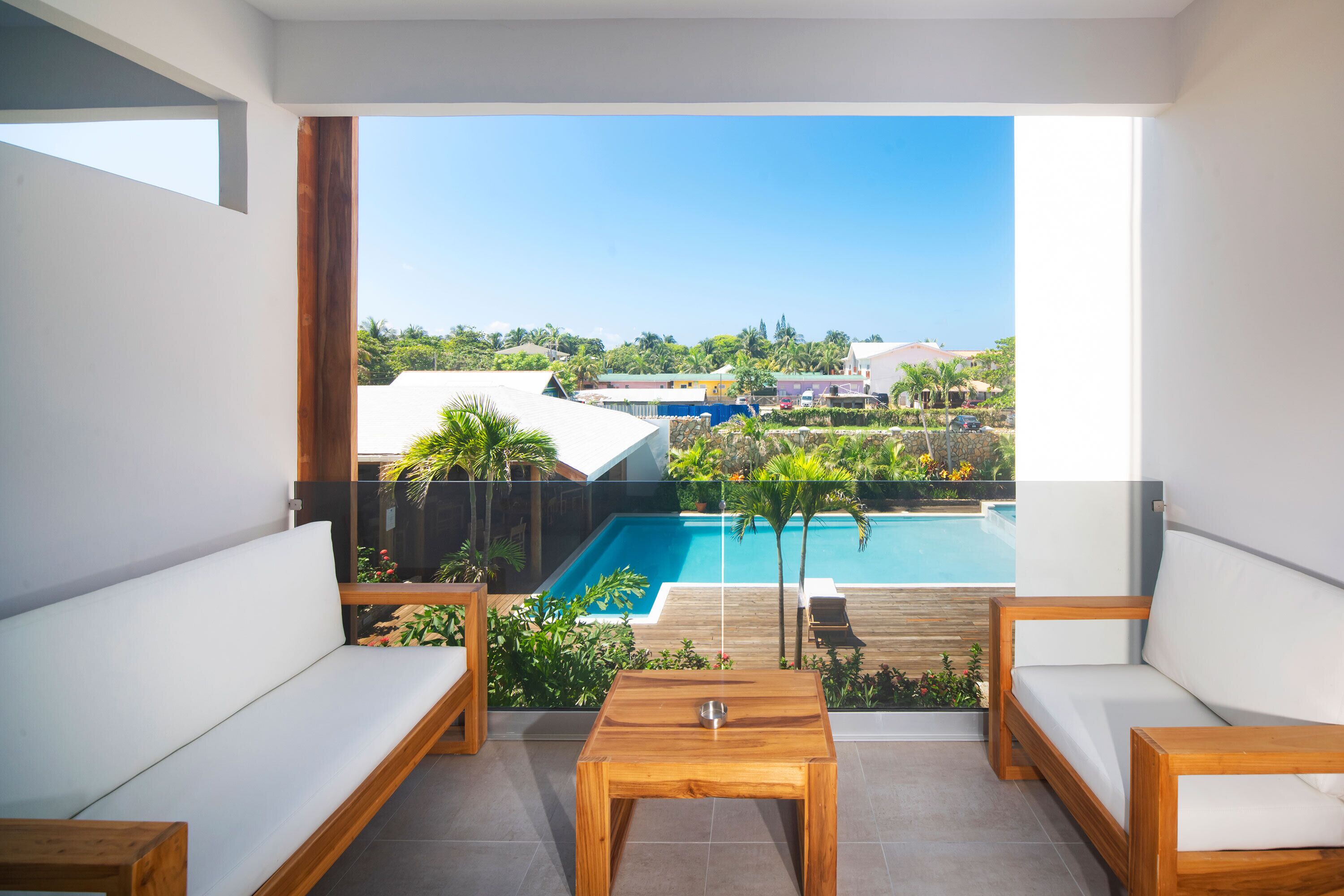 A balcony with a view of a swimming pool, wooden furniture, and a clear sky.