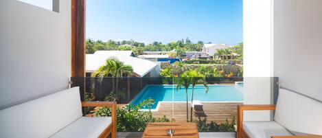 A balcony with a view of a swimming pool, wooden furniture, and a clear sky.