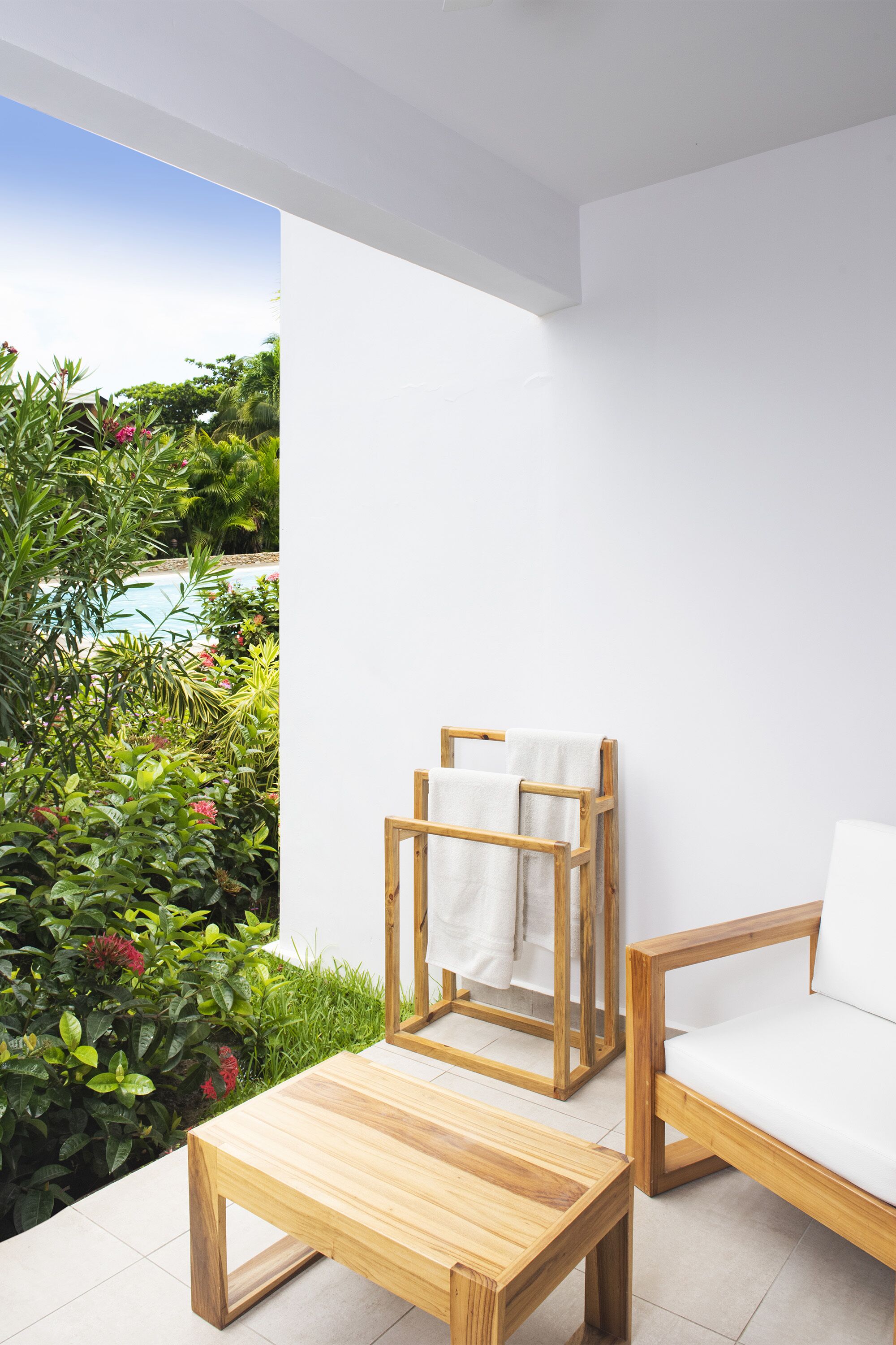 A modern outdoor seating area with a wooden table and chair, a wooden towel rack, and lush greenery in the background.