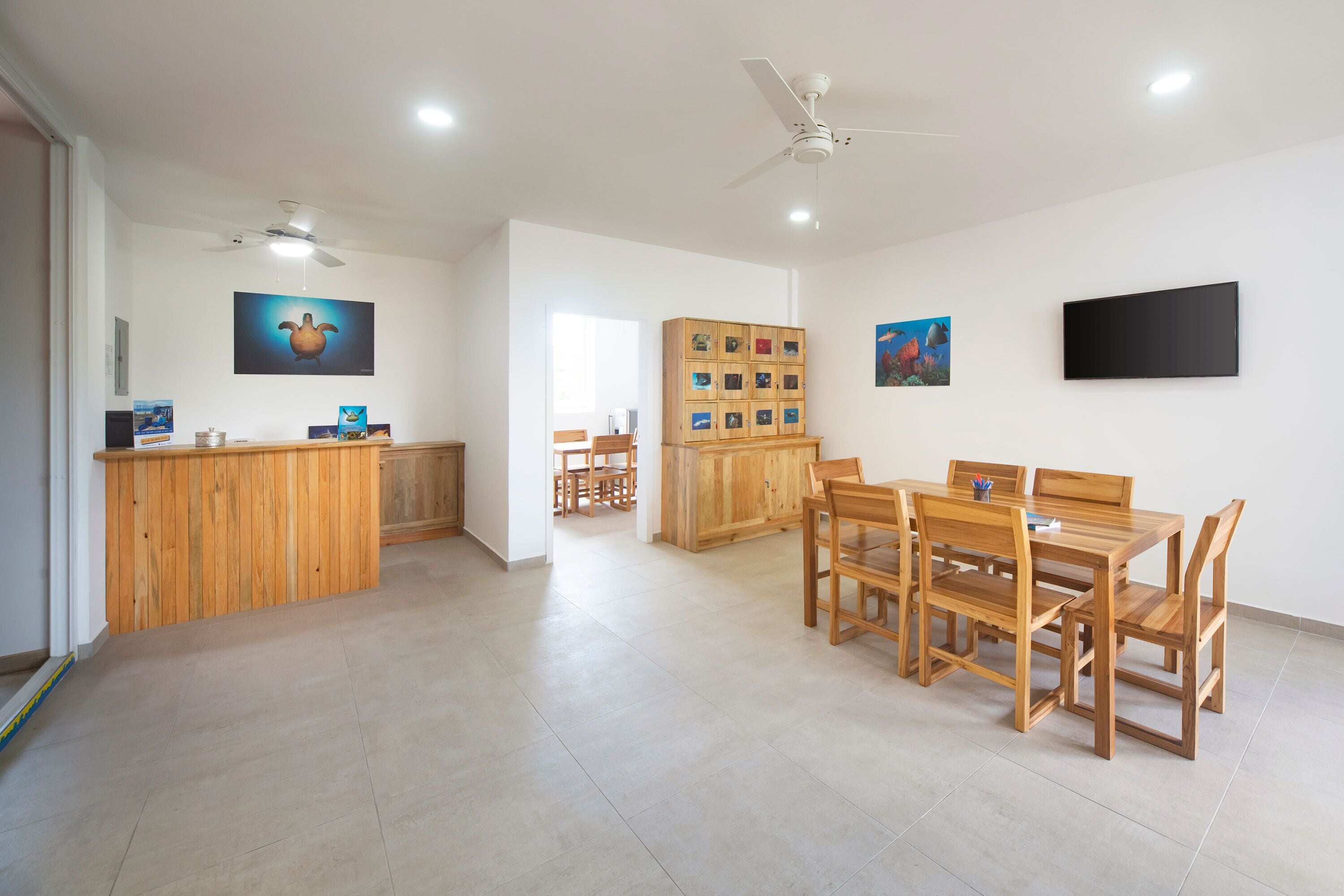 Une pièce spacieuse avec un comptoir d’accueil en bois, un coin repas comprenant une table et des chaises, un ventilateur de plafond et un téléviseur fixé au mur.