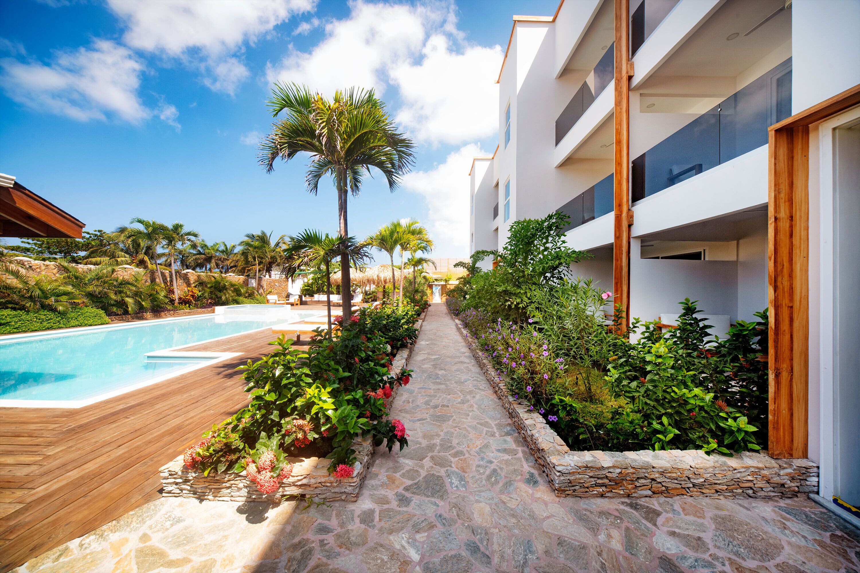 A pool area with a stone pathway, surrounded by lush greenery and a modern building with balconies.