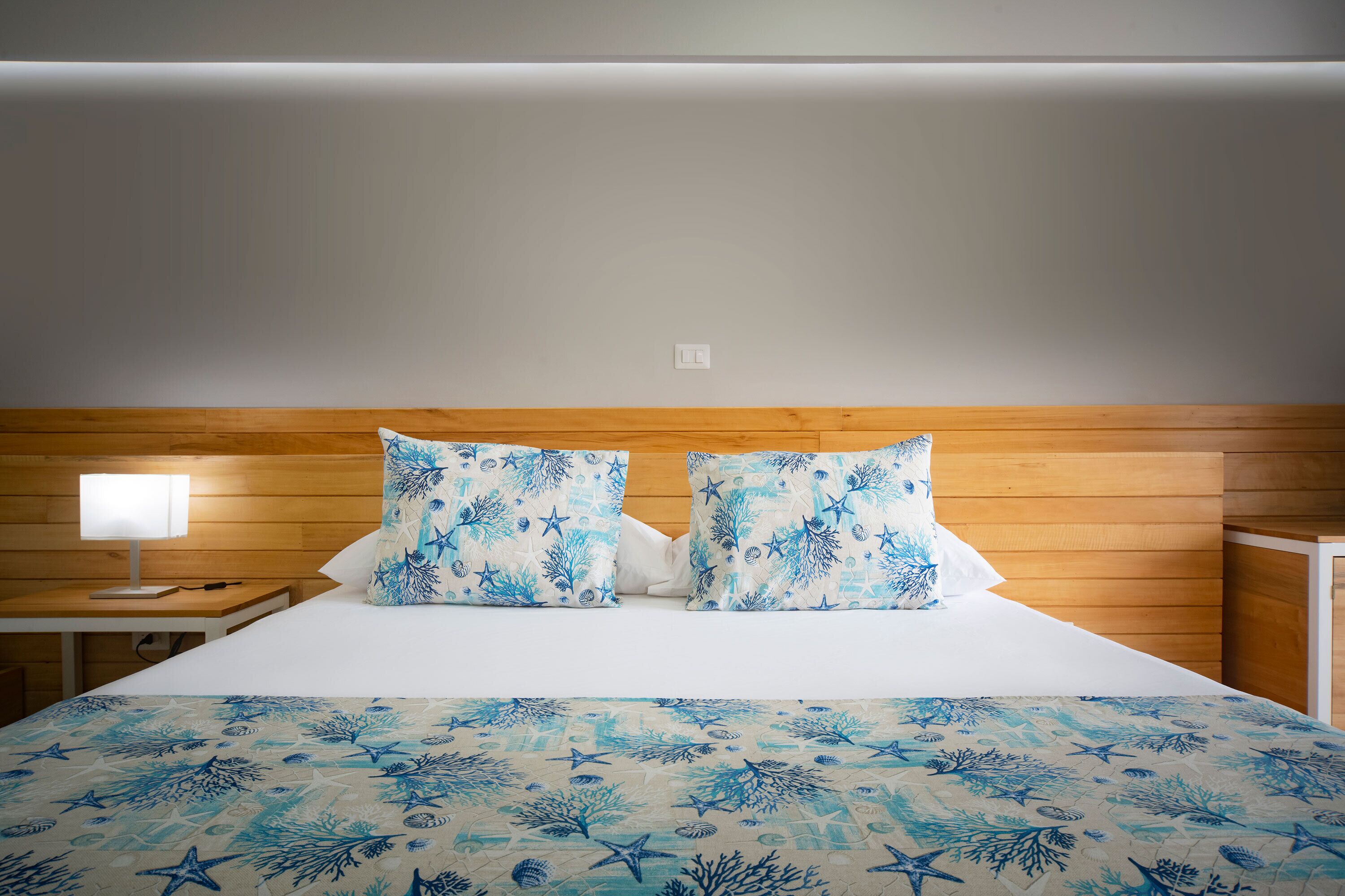 A bed with a blue and white ocean-themed bedspread and pillows, a bedside table with a lamp, and a wooden headboard.