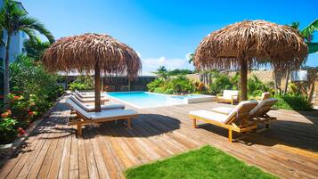 Terraza de madera con sillones de descanso y sombrillas de paja, piscina y plantas tropicales.