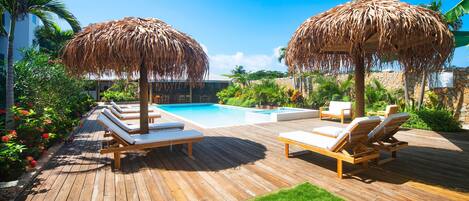 A wooden deck with lounge chairs and thatched umbrellas, a swimming pool, and tropical plants.