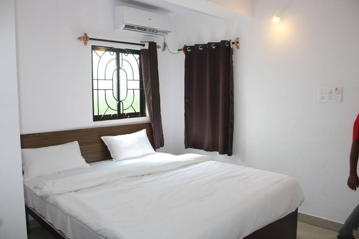 A hotel room with a bed, a window with curtains, an air conditioning unit, and a wall socket.