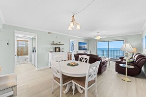 A spacious living area with a dining table, white chairs, a brown sofa, and a balcony with a view of the ocean.