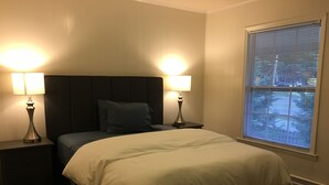 Ein ordentlich bezogenes Bett mit dunklem Kopfteil, zwei Nachttischlampen und einem Fenster mit Jalousien.