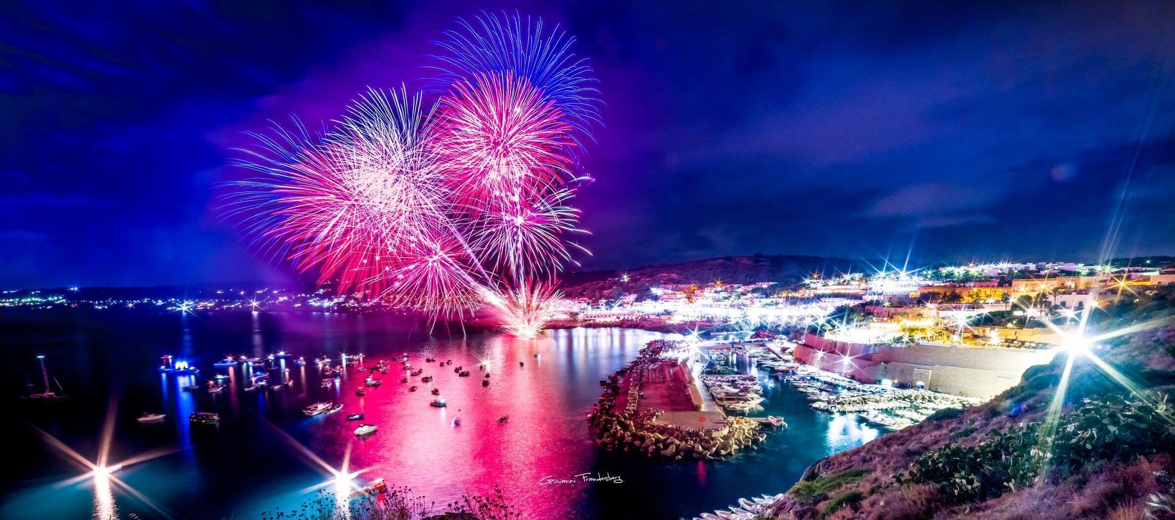 Una località costiera di notte, con fuochi d'artificio, imbarcazioni e edifici illuminati.