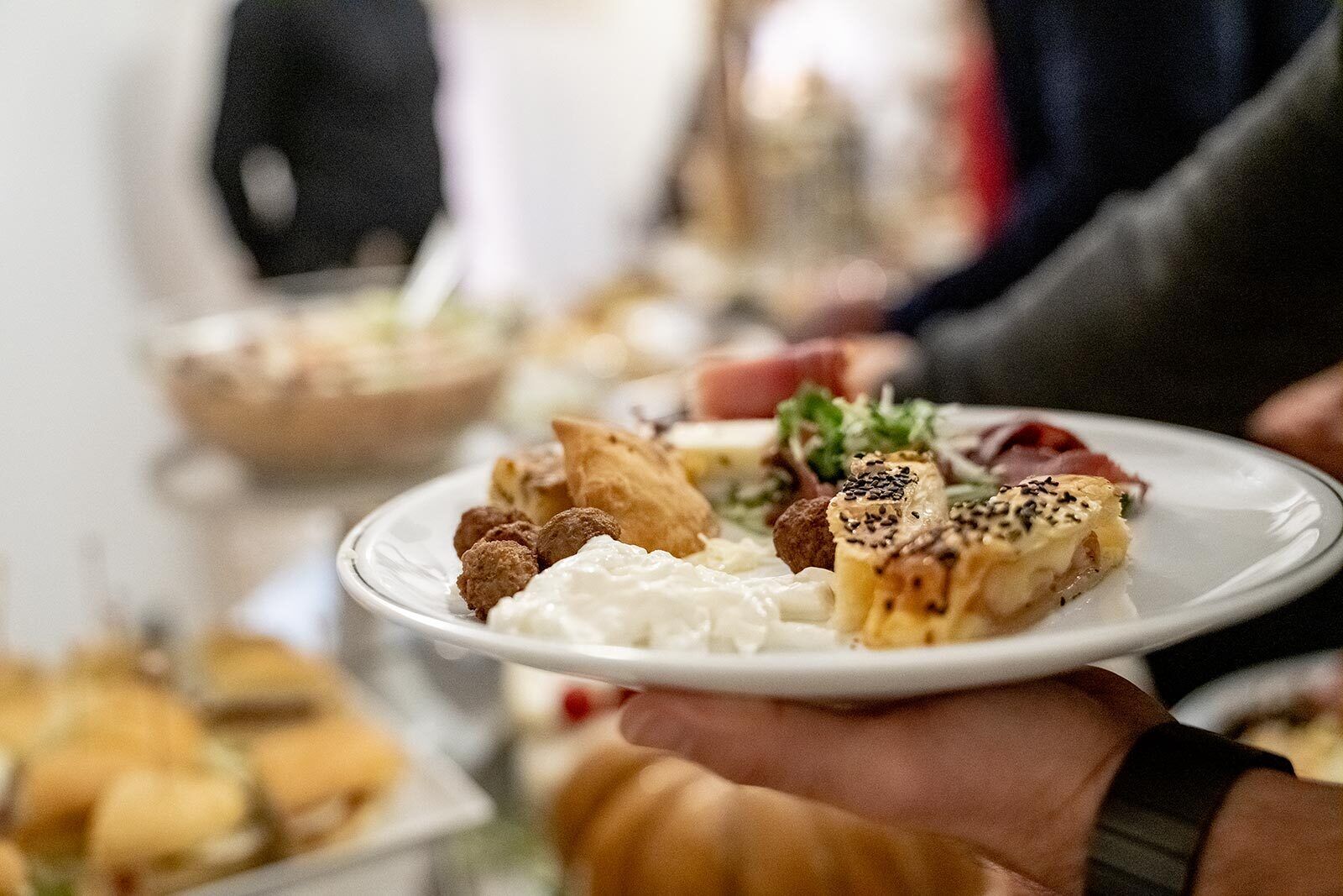 A person holding a plate with various food items, including pastries and dips, at a buffet setting.