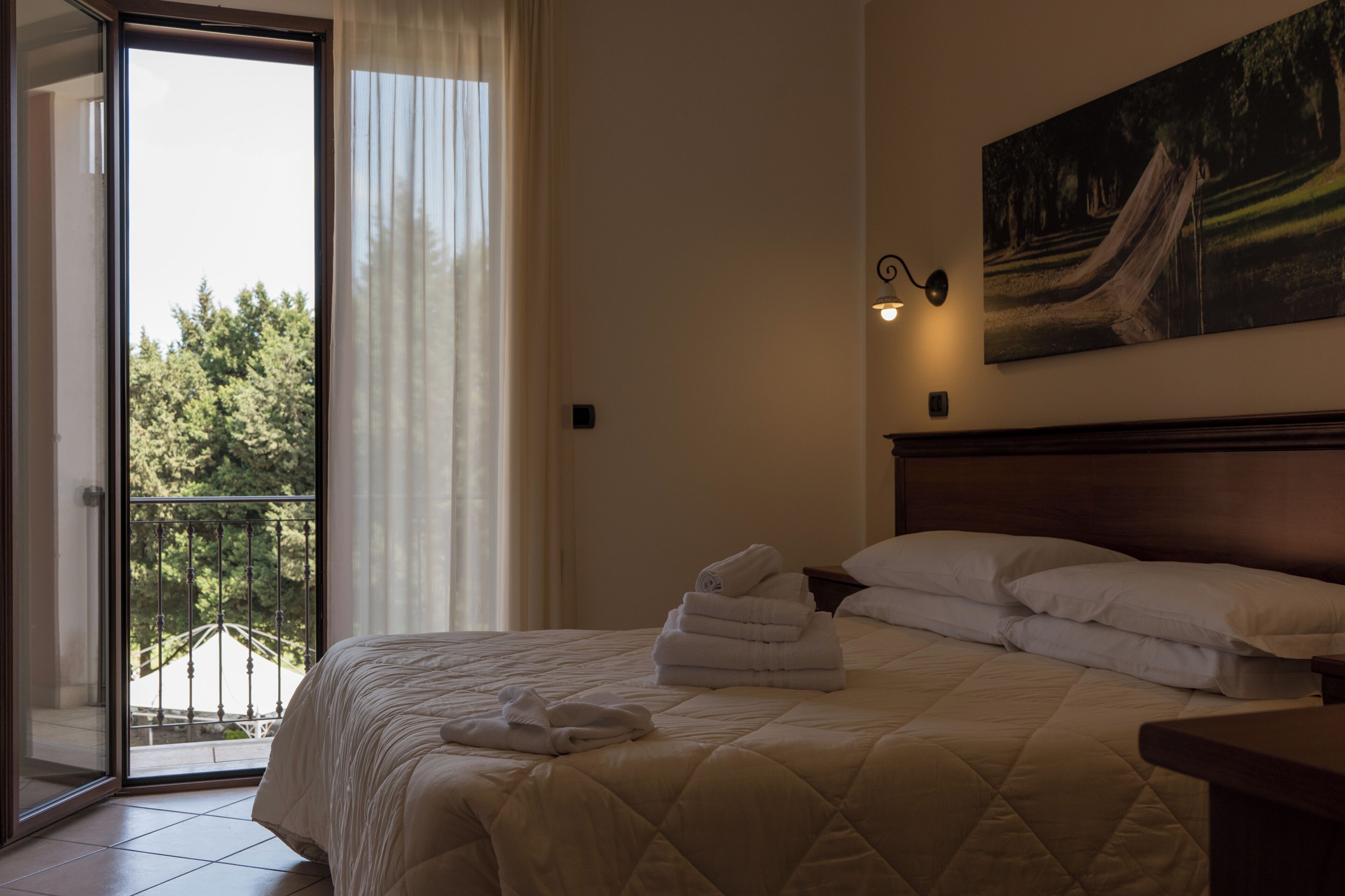 A hotel room with a bed, a painting on the wall, a balcony with a view, and a door leading outside.