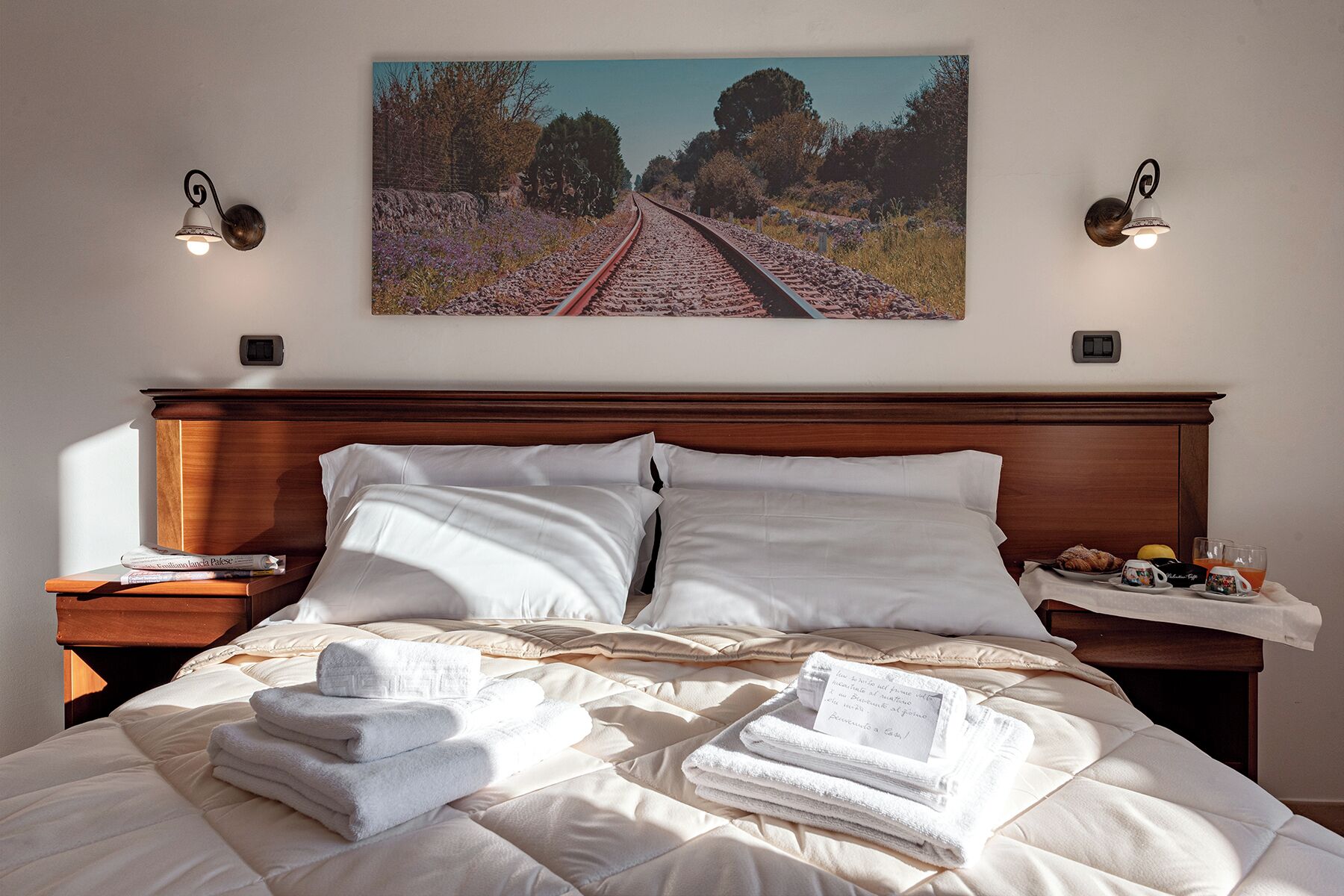 A hotel room with a bed, two bedside tables, a painting above the bed, and a nightstand with a lamp.