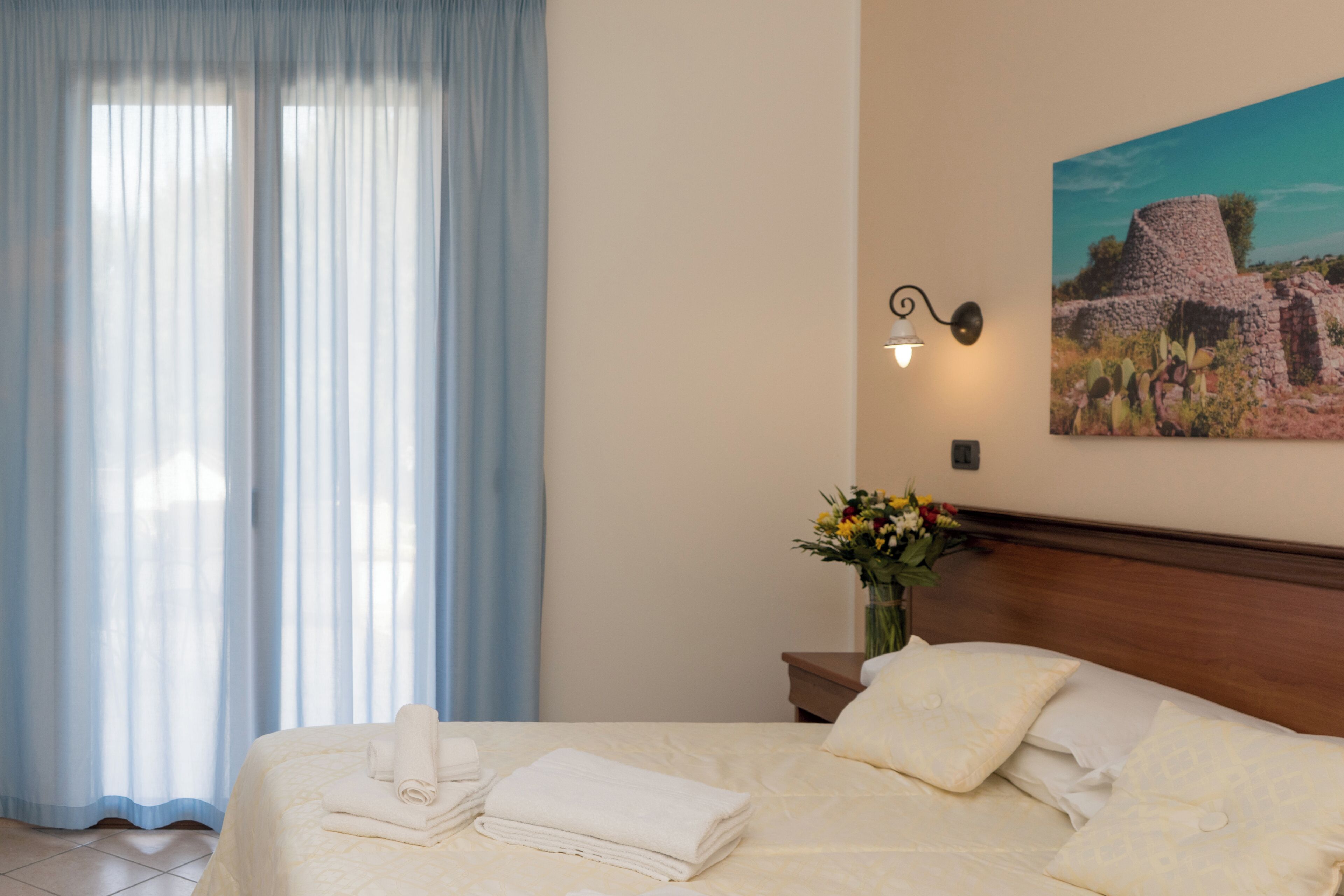 Een hotelkamer met een bed, gordijnen, een wandlamp, een vaas met bloemen en een schilderij.