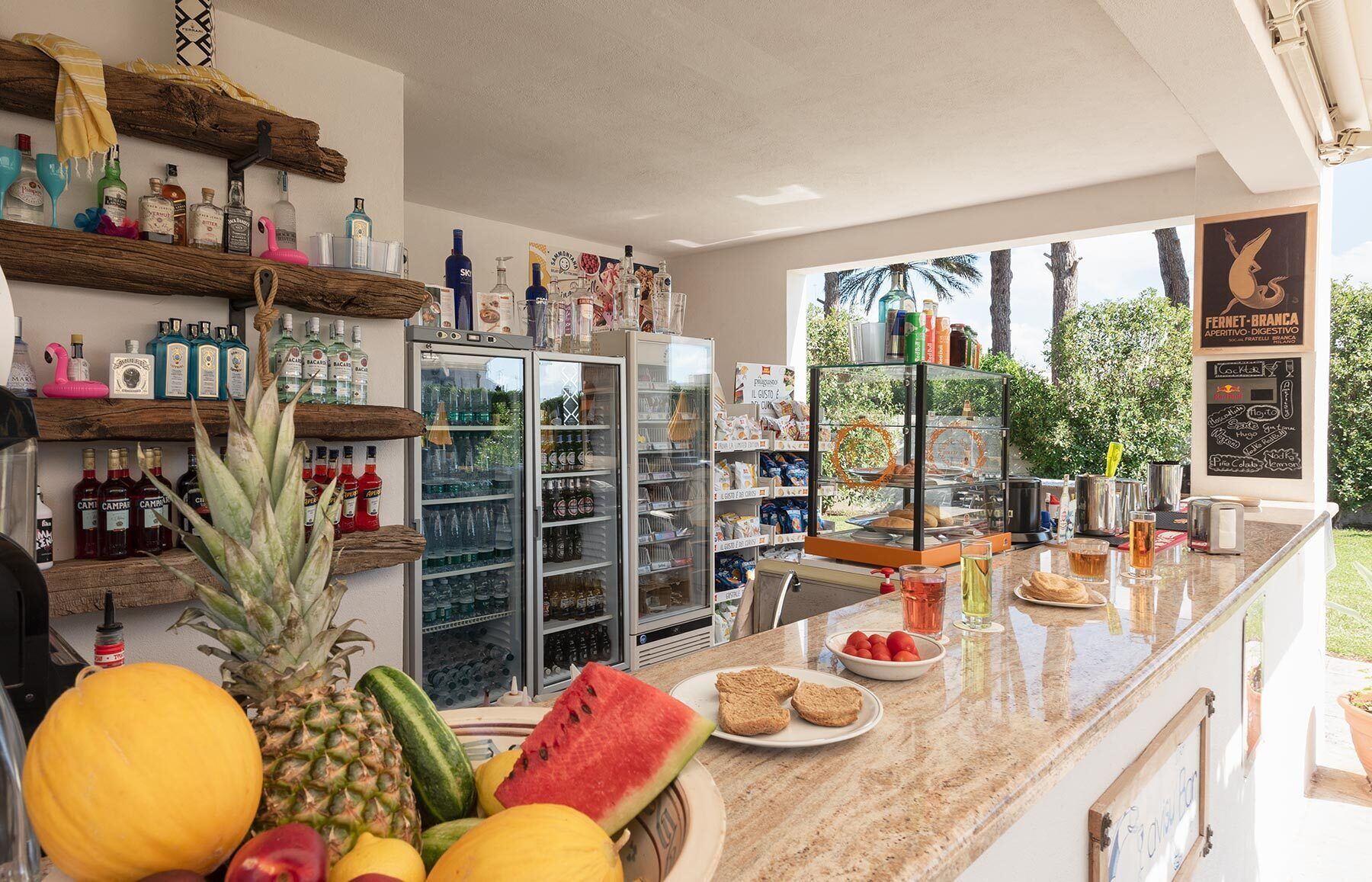 Un espace bar proposant une variété de boissons, un réfrigérateur rempli de boissons et un comptoir où l'on trouve des fruits frais et du pain.
