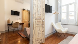 Une pièce avec un plancher en bois, un bureau avec une chaise, un téléviseur fixé au mur, un fauteuil blanc et moelleux, et une grande fenêtre.
