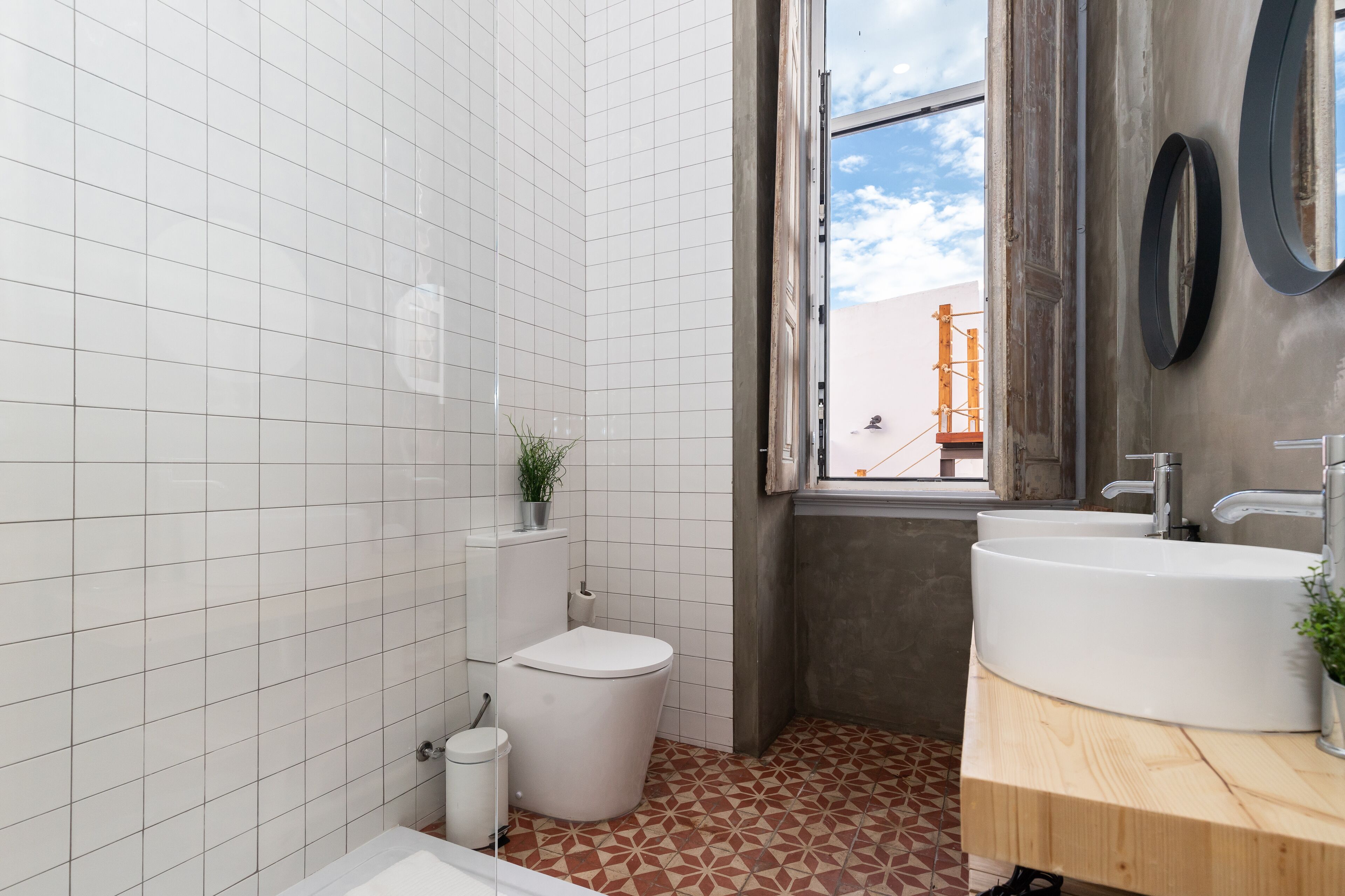 Kylpyhuoneessa on valkoisilla laatoilla päällystetyt seinät, wc-istuin, pesuallas, jossa on puinen taso, ja ikkuna, josta avautuu näkymä oranssinväriseen rakennukseen.