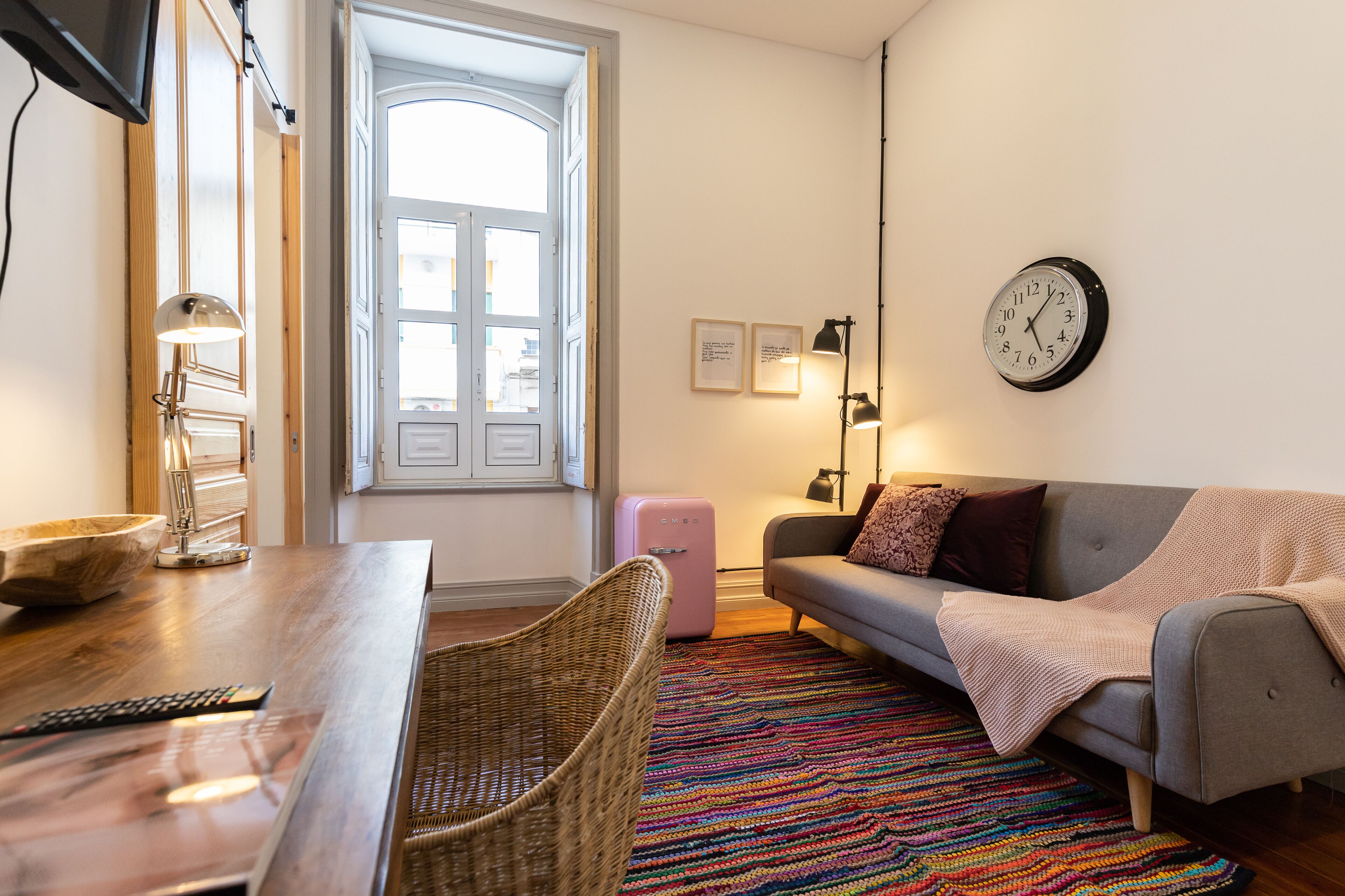 Ein gemütliches Wohnzimmer mit Holztisch, Korbstuhl, grauem Sofa, farbenfrohem Teppich, rosa Koffer und einer Wanduhr.