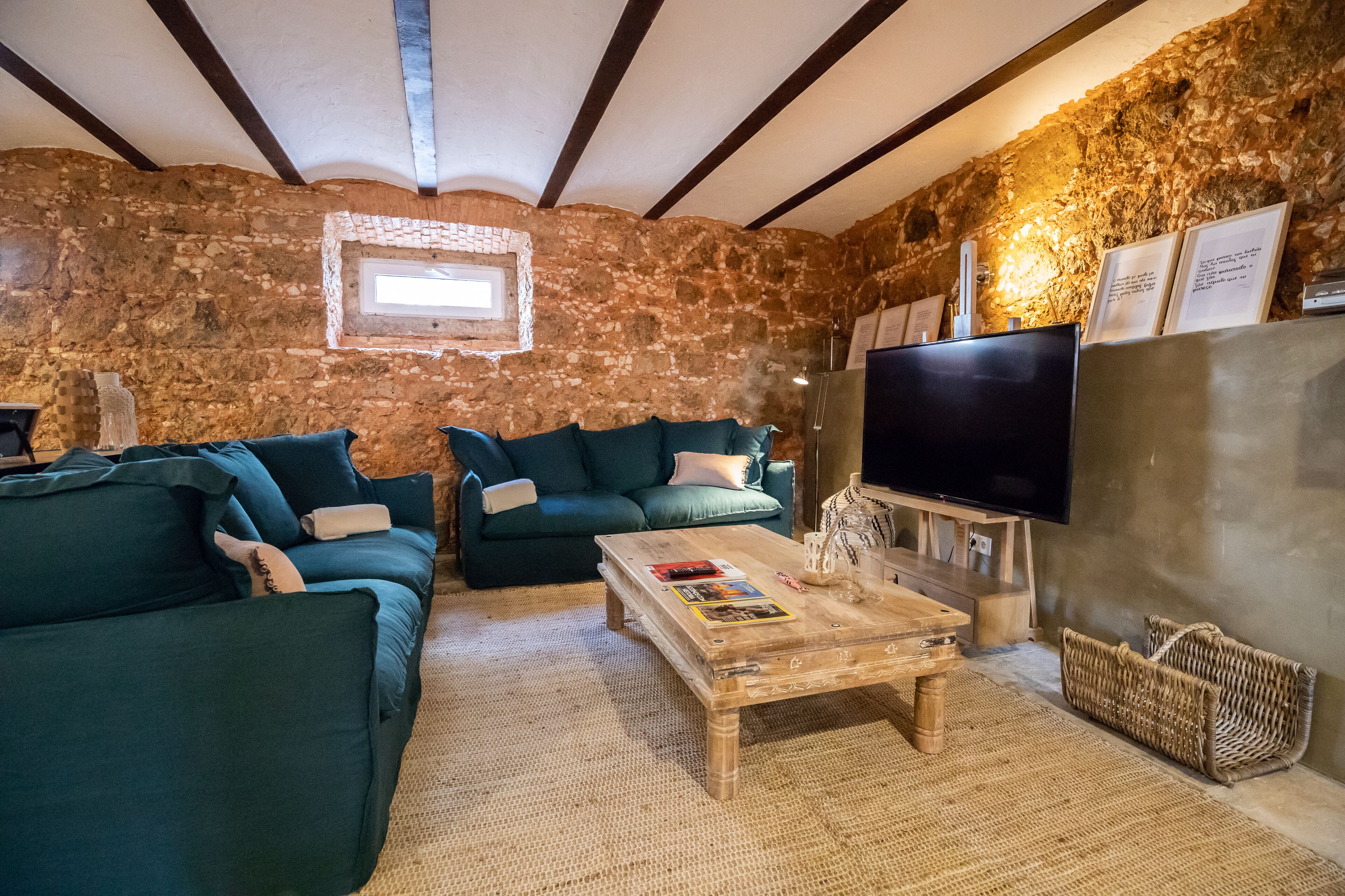 Un saló rústic amb parets de pedra, sostre de fusta, un televisor de pantalla plana gran i dos sofàs verds.