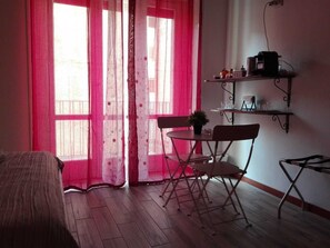 A room with a bed, a small table with chairs, a potted plant, and a shelf with items.