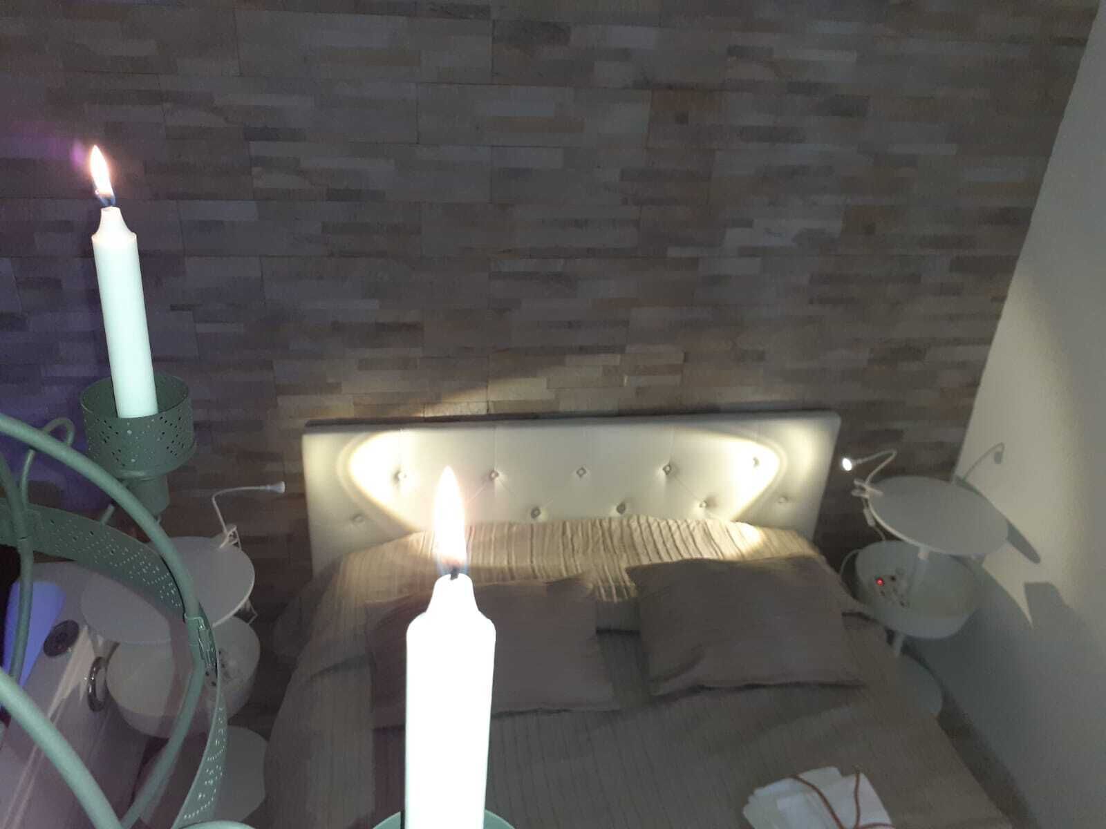 Quarto com cabeceira estofada, mesa de cabeceira com abajur e uma vela acesa no criado-mudo.