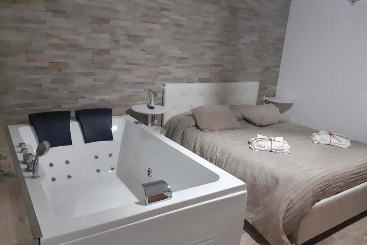 Ein modernes Schlafzimmer mit einer großen Badewanne, einem Bett mit beigefarbener Bettwäsche und einem kleinen Nachttisch mit einer Lampe.