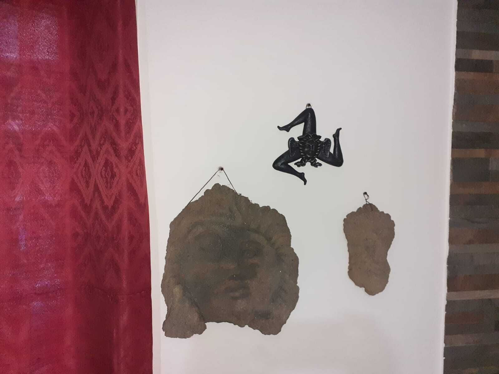 A wall with a red patterned curtain on the left and a white wall with three hanging objects: a black sculpture, a large textured stone, and a smaller stone.