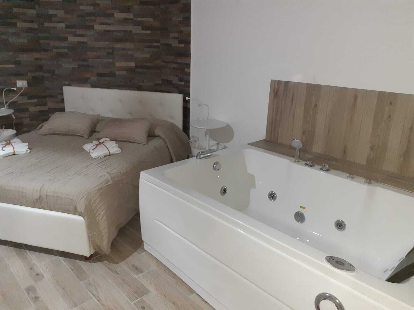 A modern bedroom with a large bed, a wooden headboard, a white bathtub, and a small bedside table with a lamp.