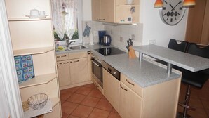 A compact kitchen with a sink, stove, and countertop. There is a clock on the wall and a window with a curtain.