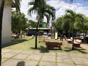 Uma área externa ensolarada com baloiços, escorregas, bancos, palmeiras e um caminho pavimentado.