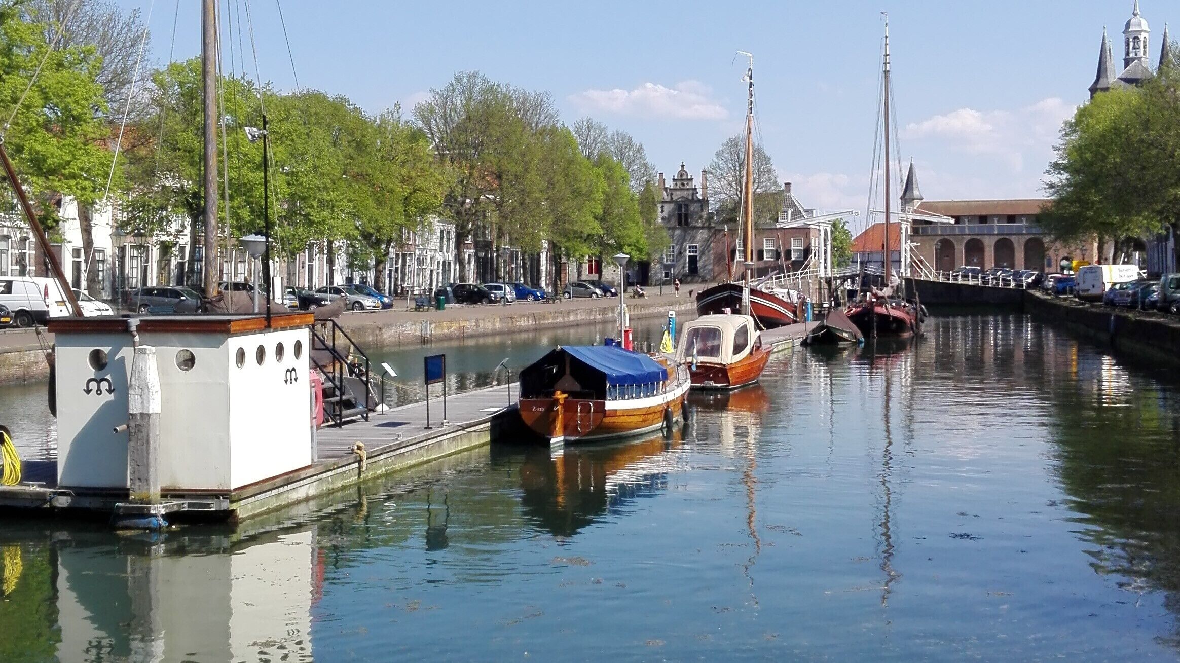 Een kanaal met aangemeerde boten, een klein houten bootje met een blauwe zeilzeil, en een wit gebouw met een bord.