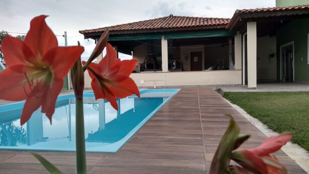 A swimming pool with a wooden deck, a green-roofed building, and red flowers in the foreground. - Family house, with pool views of the mountains 7 minut from the center Penedo RJ (Itatiaia)