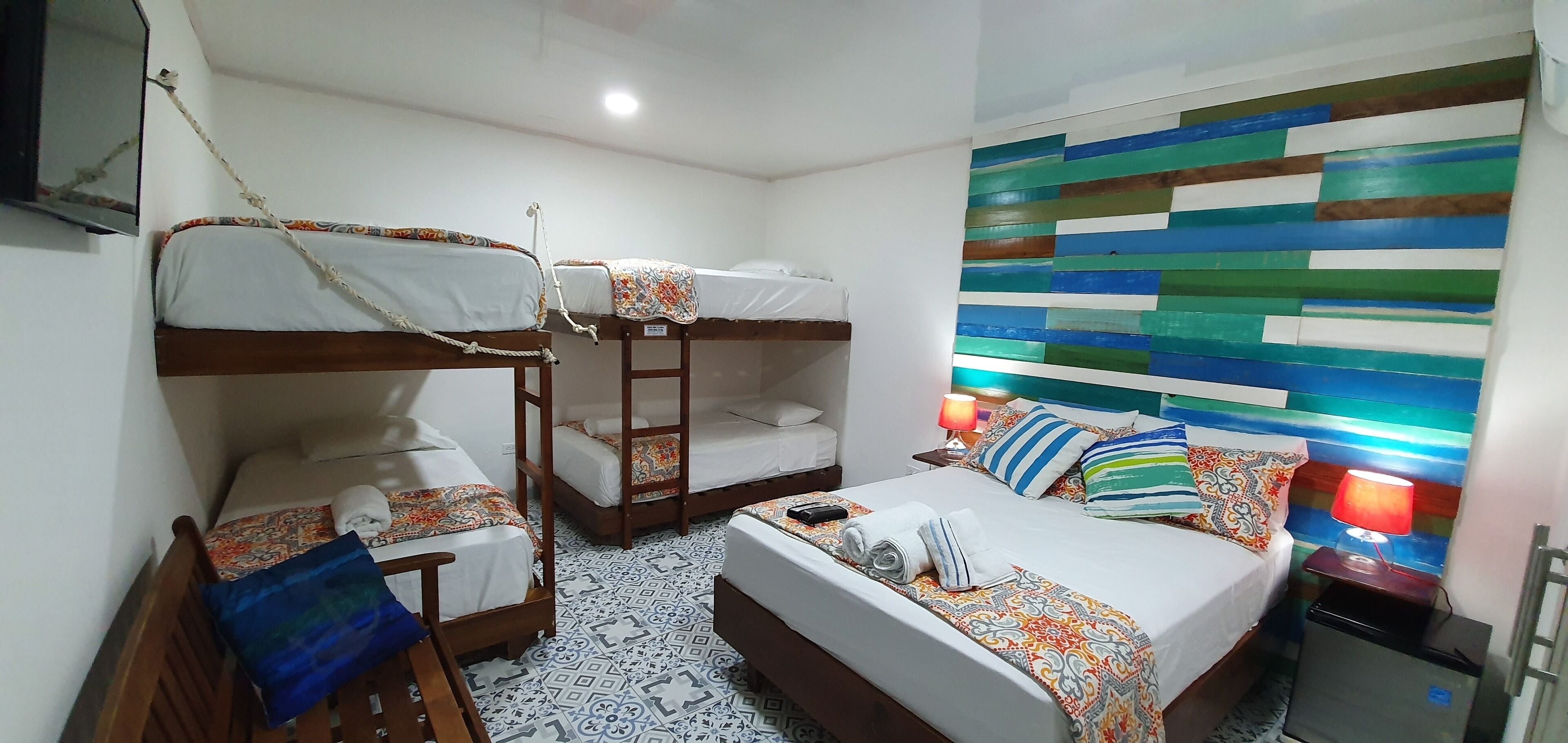 Une chambre d’hôtel avec des lits superposés, un lit recouvert d’une couvre-lit à motifs, une table de chevet avec une lampe et un sol carrelé.