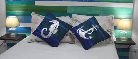 Una cama con una colcha blanca adornada con criaturas marinas, flanqueada por dos mesitas de noche con lámparas. El cabecero está pintado con franjas horizontales en varios tonos de azul y verde.
