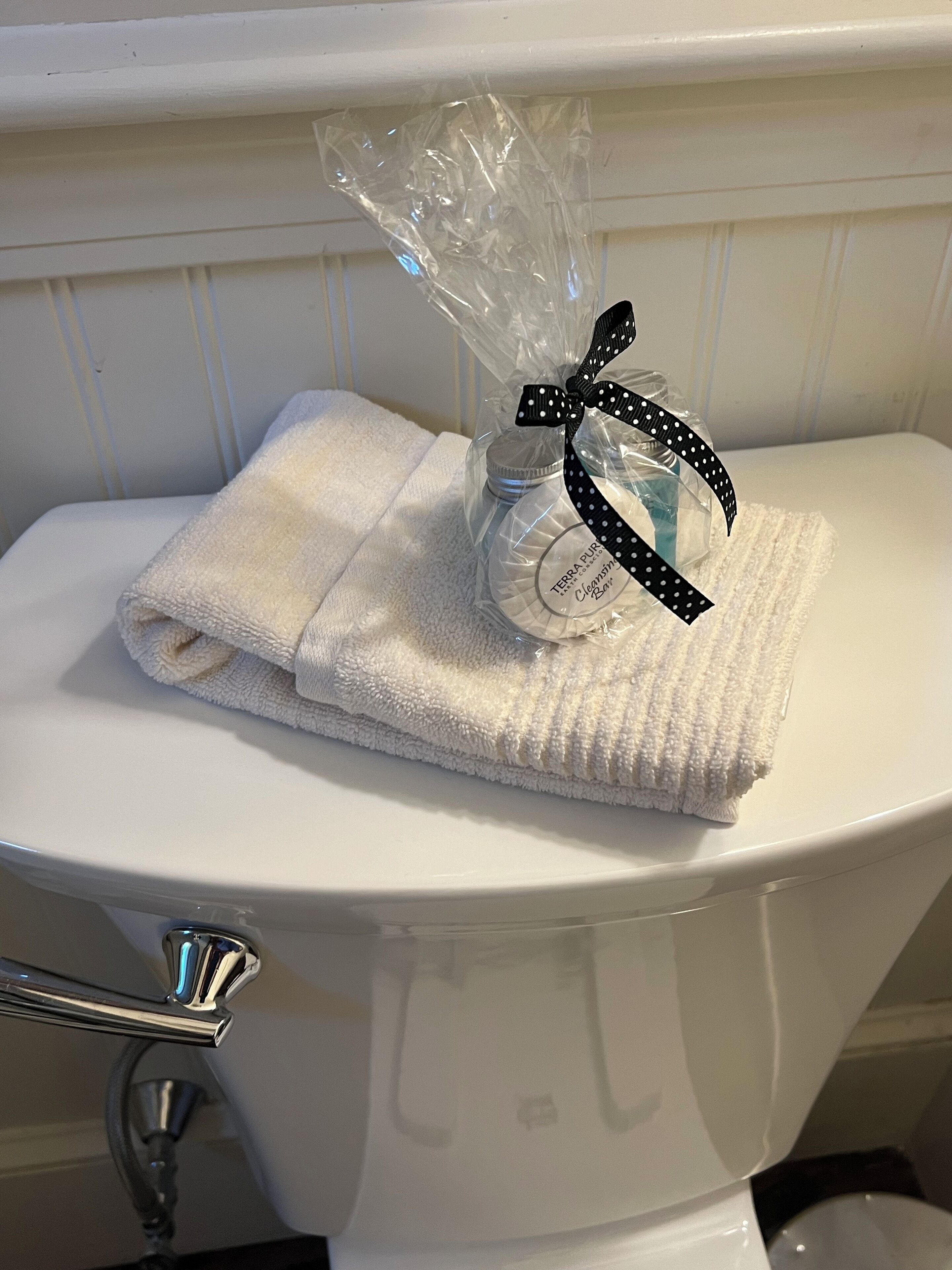 A toilet with a folded white towel on top and a plastic bag with a bottle and a black ribbon.