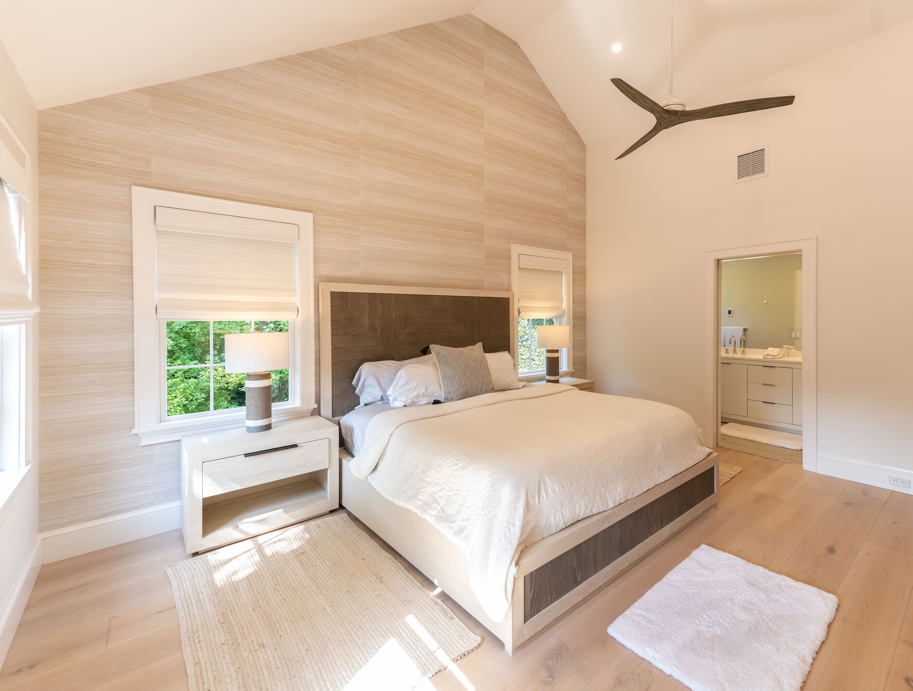 A bedroom with a large bed, bedside tables, a nightstand, a ceiling fan, a window with blinds, and a bathroom visible through an open door.