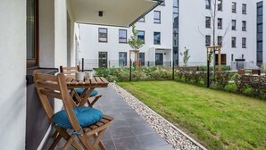 A modern outdoor seating area with wooden chairs and a table, a small garden with a fence, and a building with multiple windows and balconies in the background.